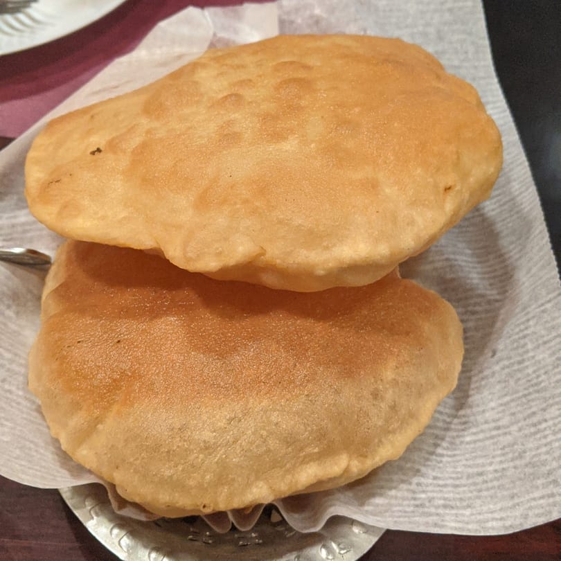 Two golden-brown, puffy Indian puri breads served on a white paper liner in a metal basket.